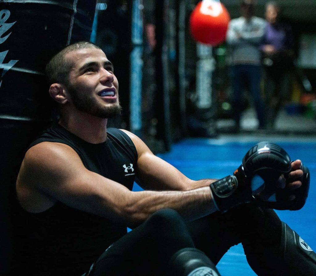 MMA fighter Muhammad Mokaev resting against cage in Under Armour gear during intense training Wallpaper