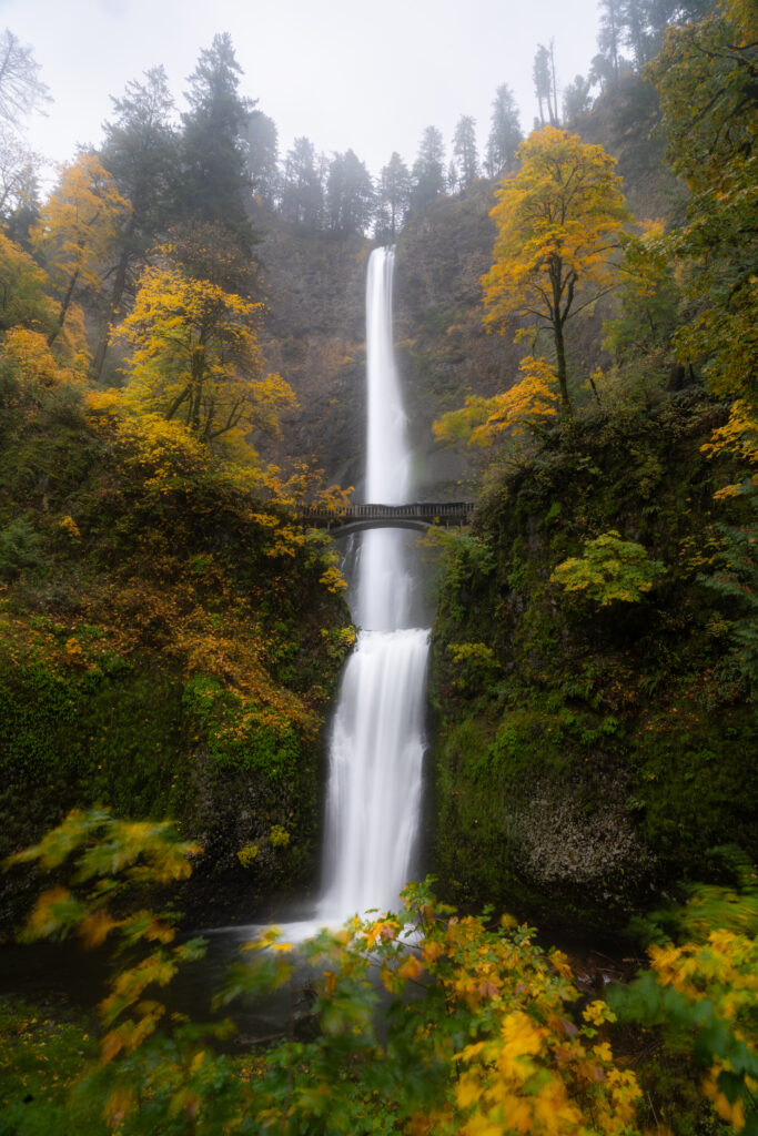 Tall multi-tier waterfall with vibrant autumn trees and arched bridge in misty forest wallpaper