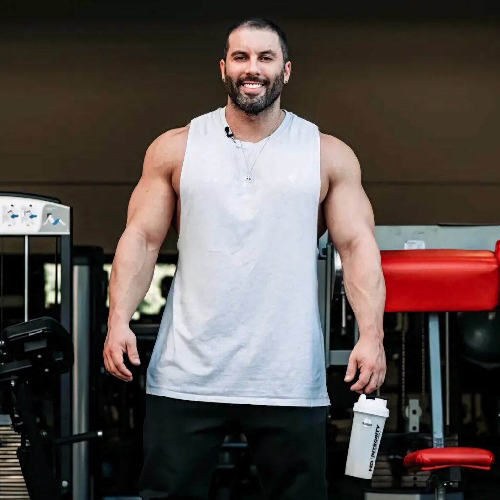 Muscular bodybuilder in white tank top showing strong arms confident smile gym equipment background Wallpaper