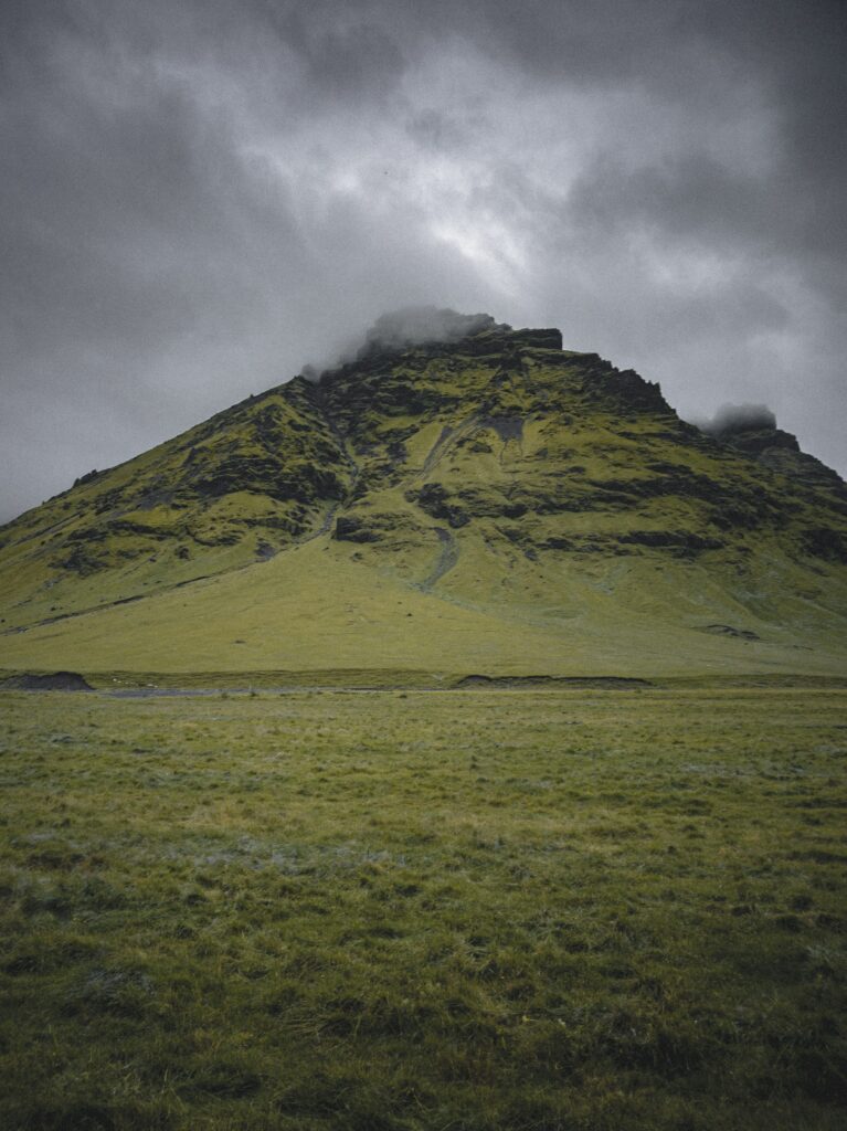 Dramatic mountain peak with lush green grass field and foggy summit under moody cloudy sky Wallpaper