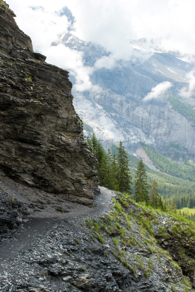 Narrow mountain trail along rocky cliffs with evergreen trees and misty mountains background Wallpaper