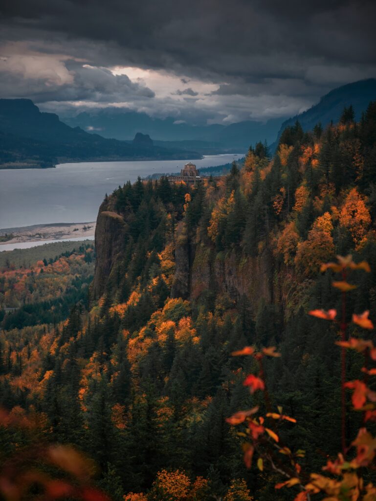 Dramatic autumn cliffs with vibrant forest river and solitary building under moody skies wallpaper