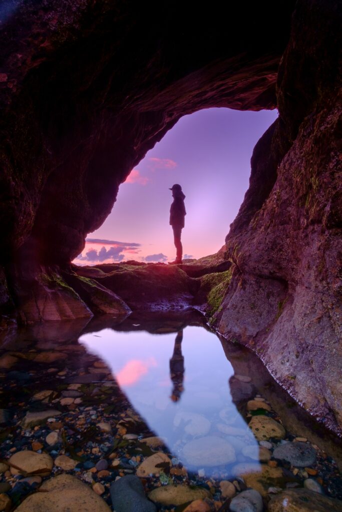 Silhouette of lone figure at cave entrance with sunset sky and clear water reflection wallpaper background