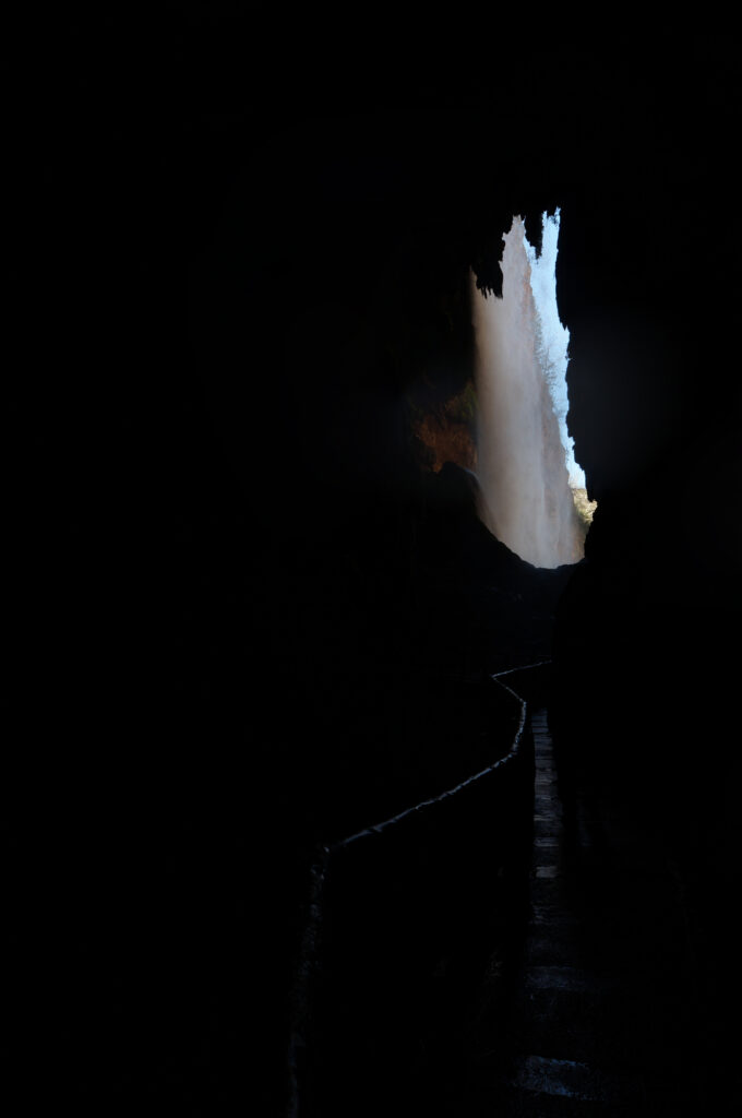 Dark cave with winding path leading to a bright majestic waterfall illuminated by natural light Wallpaper