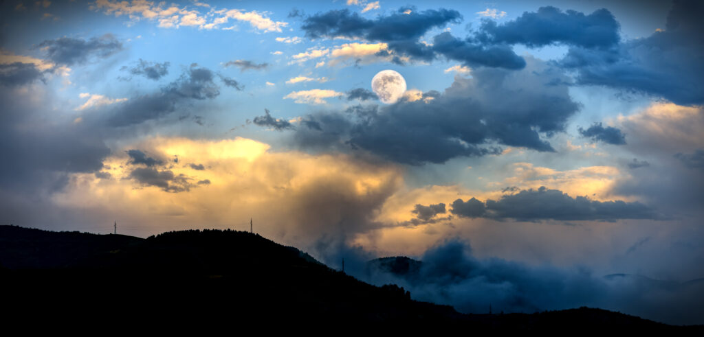 Full moon illuminates swirling colorful clouds over foggy hills creating atmospheric wallpaper background