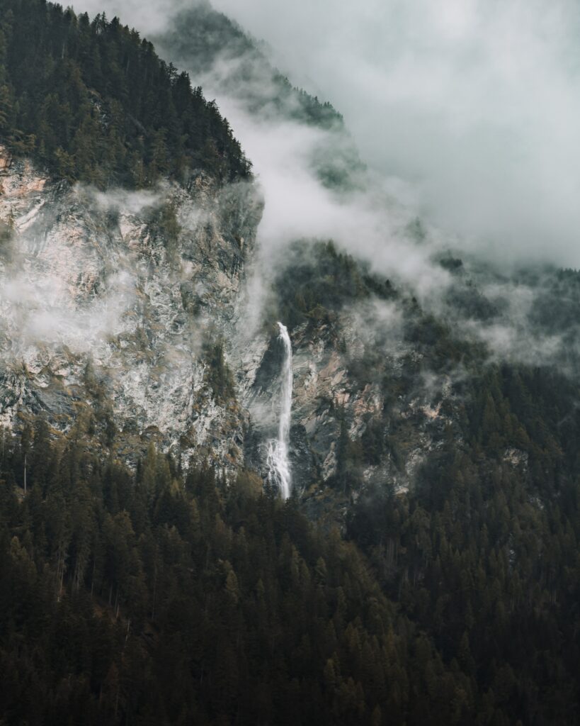 Dramatic mountain landscape with rugged rocks dense spruce trees and misty waterfall background Wallpaper