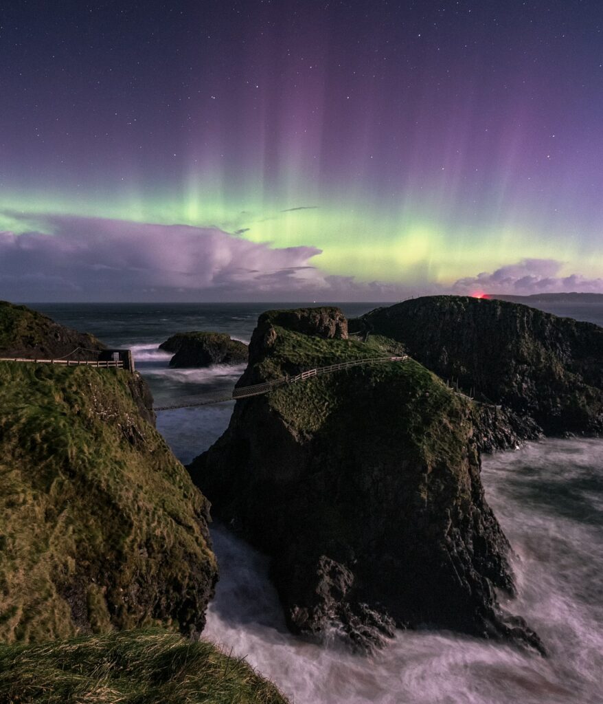 Northern lights illuminate jagged coastal cliffs and wooden bridge over churning sea at night Wallpaper