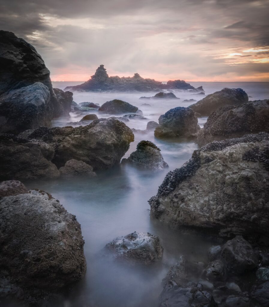 Mystical ocean landscape with rugged rocks and fog under a dramatic sunset sky wallpaper background