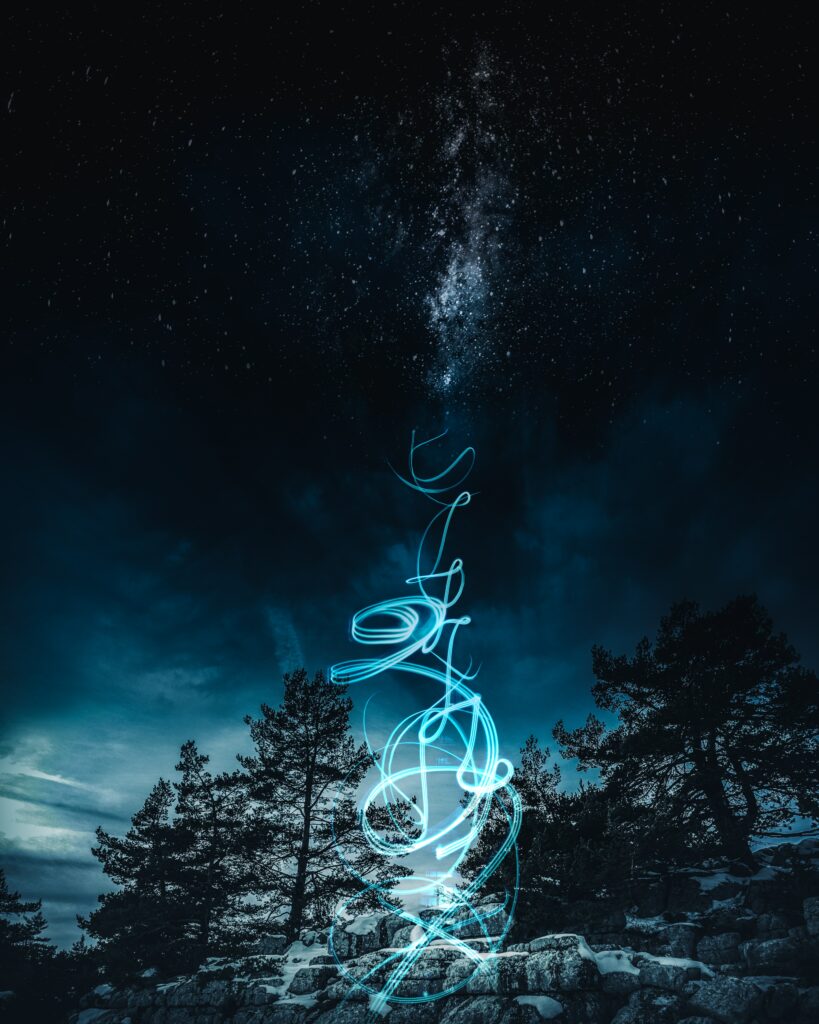 Starry night sky with silhouetted trees and glowing blue light art over rocky terrain wallpaper