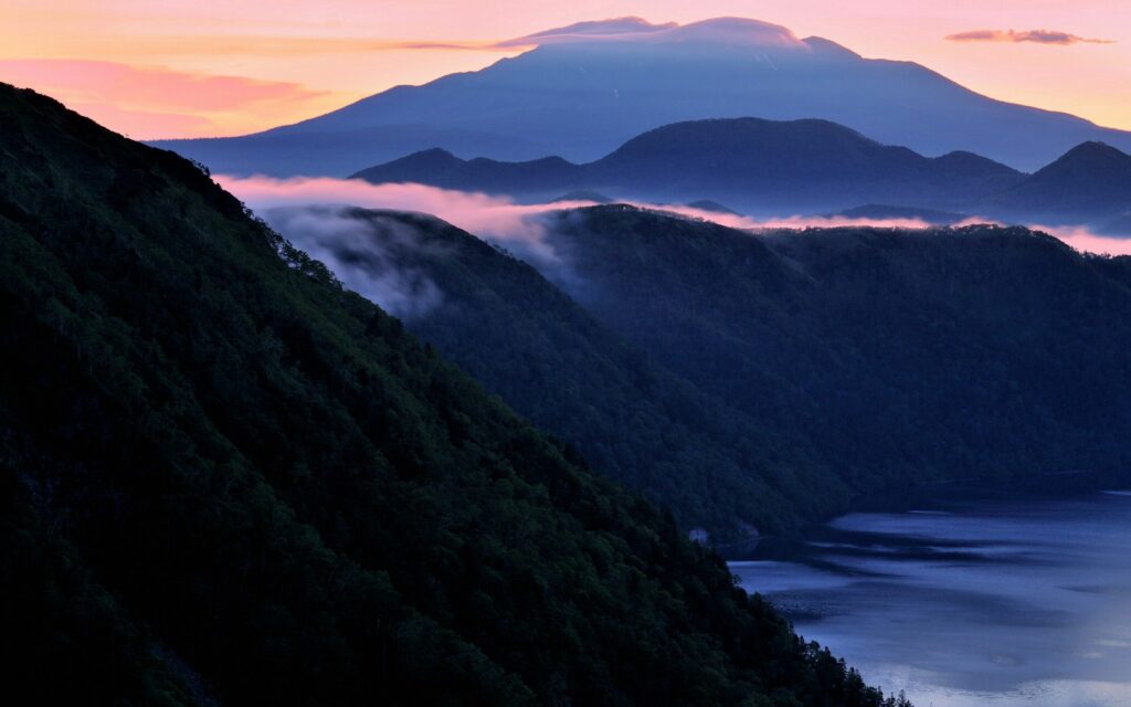 Mist-covered forested mountains at twilight by calm sea creating tranquil mystical night scene wallpaper