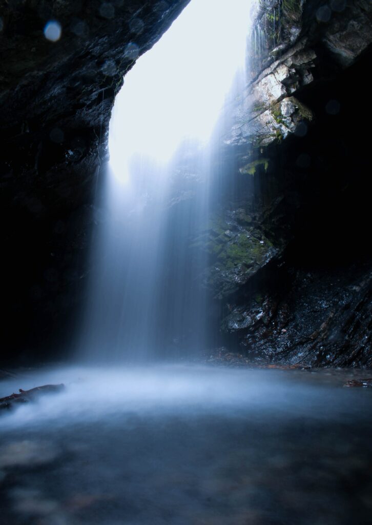 Mystical waterfall flowing through cave opening with sunlight mist and dark stone textures wallpaper