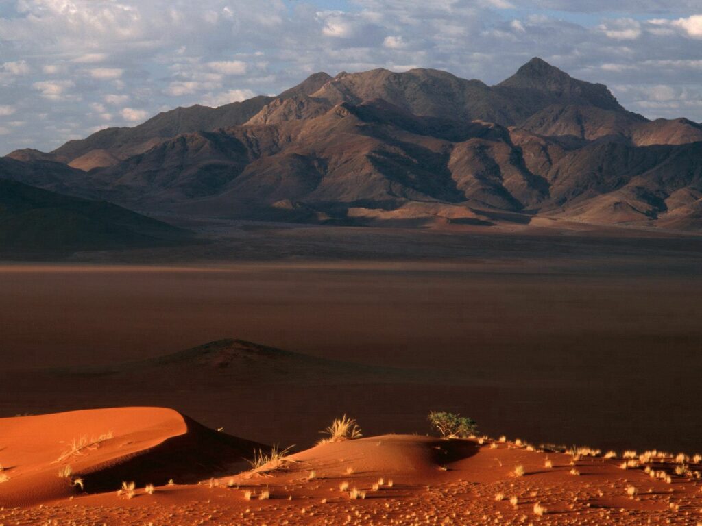 Namibia desert wallpaper with red sand dunes and sunlit mountains under a cloudy sky