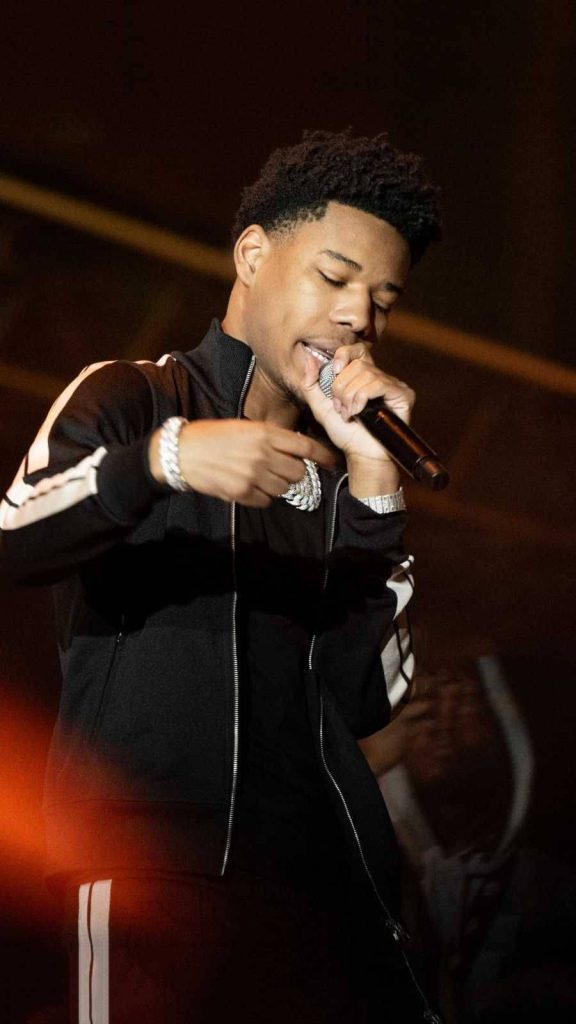 Nardo Wick performing live in black striped jacket and iced jewelry on dark moody stage Wallpaper