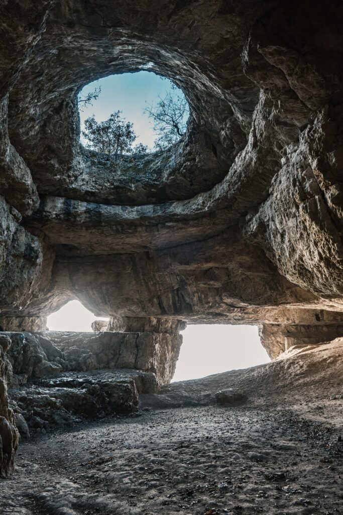Natural cave interior with rugged stone formations lit by soft daylight through openings wallpaper