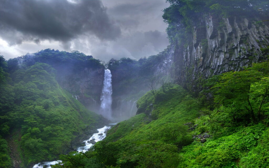 A Stunning HD Wallpaper of a Green Tree Landscape with a Majestic Waterfall, River and Forest Peeking Through the Clouds