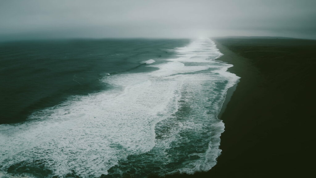 Aerial view of moody coastline with white waves and dark sand under misty sky beach wallpaper