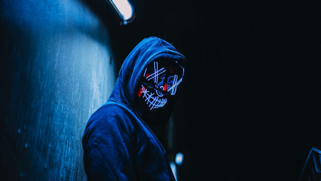 Unveiled Anonymity: High Definition Image of a Mysterious Figure Balancing the Hacker Aesthetic, Adorning Neon Mask and Hoodie Jacket Against a Blue-effect Wall Wallpaper