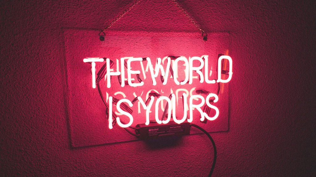 Neon sign reading THE WORLD IS YOURS glowing bright red on dark textured wall background Wallpaper