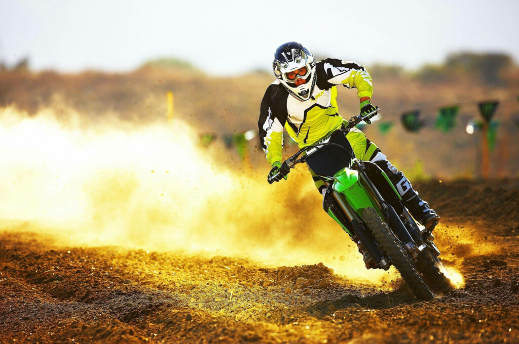 Dirt bike rider in neon gear racing on rugged track kicking golden dust bright sunny motocross action Wallpaper
