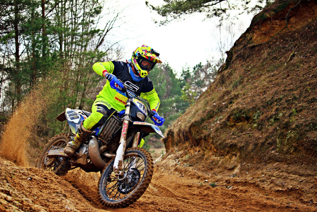 Dirt bike rider in neon yellow gear speeds on rugged trail kicking up dirt with forest background Wallpaper