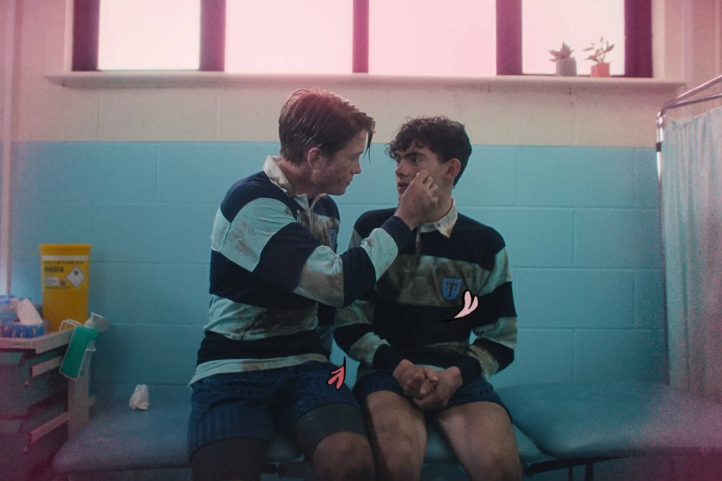 Nick and Charlie from Heartstopper in rugby uniforms sharing a tender moment in medical room Wallpaper