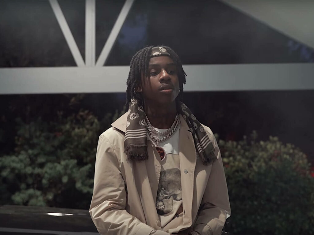 Polo G in beige trench coat and skull bandana with iced chains against dark foliage wallpaper