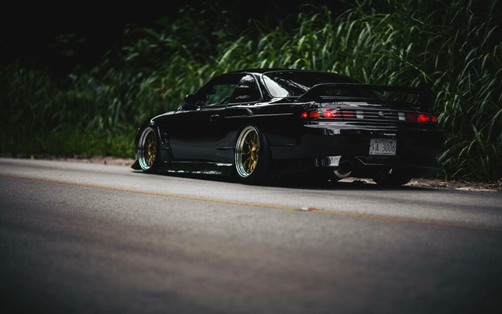 Rear view of Nissan Silvia S14 with custom wheels parked low on street surrounded by greenery Wallpaper
