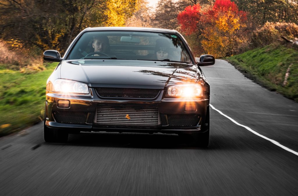 Nissan Skyline GT-R R34 black JDM car driving on autumn road with vibrant fall foliage background Wallpaper
