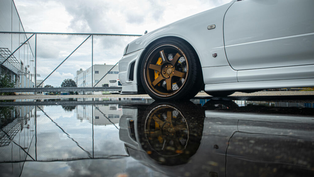 Close-up of Nissan Skyline GTR R34 bronze rim wheel by reflective puddle urban sporty design wallpaper