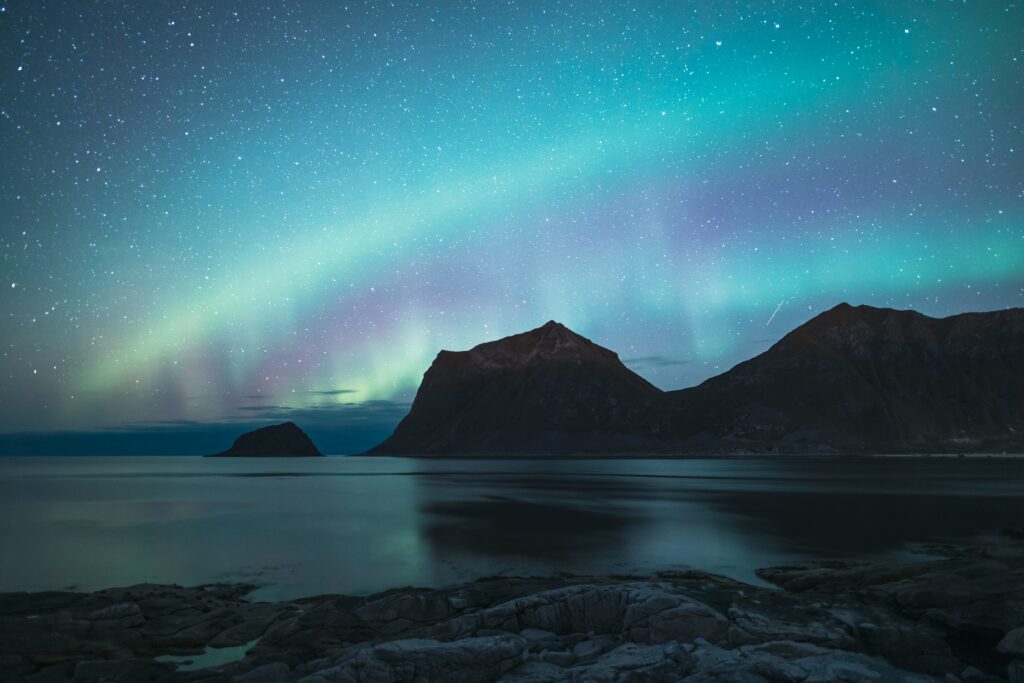 Vibrant northern lights over rugged sea rocks and mountains reflecting on calm waters wallpaper background