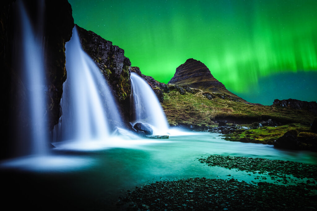 Cascading waterfall into tranquil pool with pebbles under northern lights and mountain silhouette Wallpaper