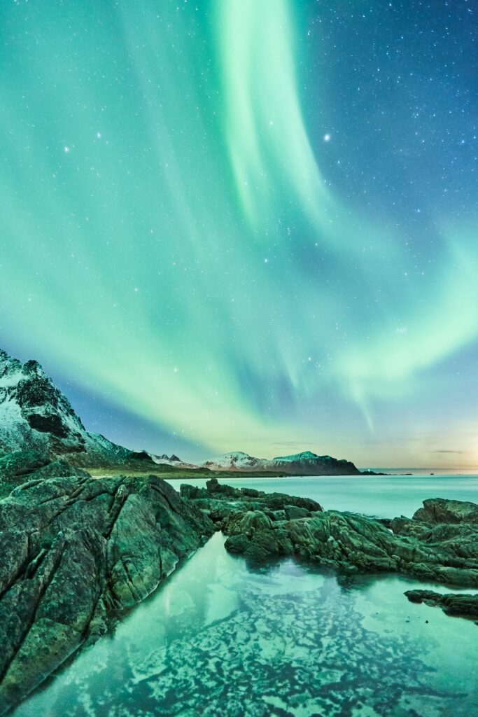 Northern lights over starry sky reflected in lake with snowy mountains and rocky shore wallpaper
