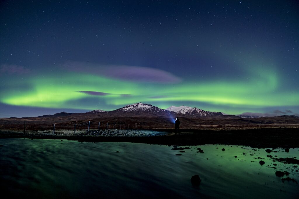 Northern lights glow green over snow mountains with lone figure by river under starry sky Wallpaper
