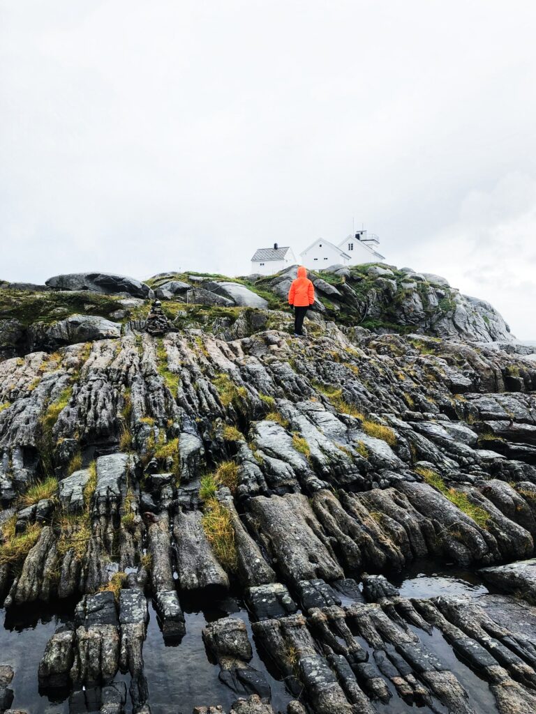 Man in orange jacket on rugged rocks under cloudy sky near white house Norwegian winter landscape Wallpaper