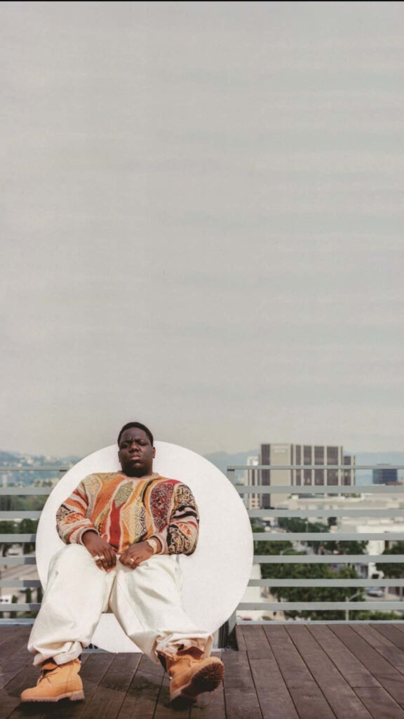 Notorious B.I.G. wearing colorful sweater and white pants against vibrant cityscape background Wallpaper