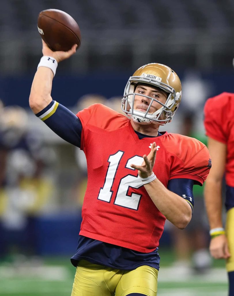 Notre Dame quarterback Ian Book in gold helmet throwing football in red practice jersey wallpaper