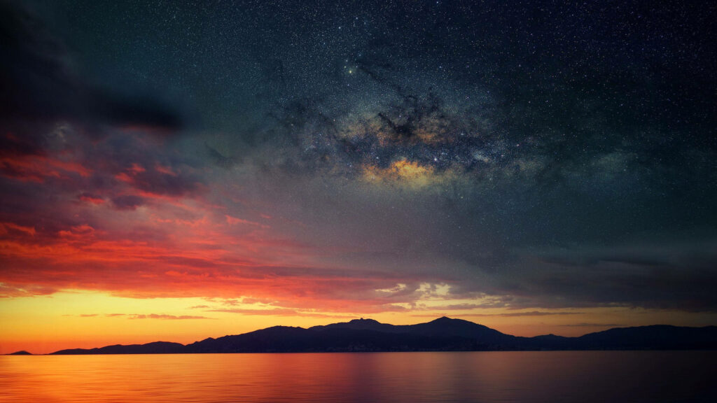 Ocean sunset reflecting golden hues beneath a vibrant starry galaxy sky digital wallpaper background