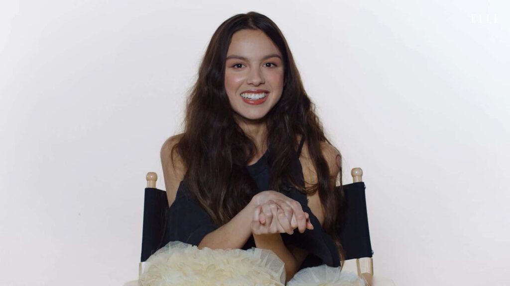 Olivia Rodrigo Smiling Warmly on Chair - Relaxed Wallpaper Background