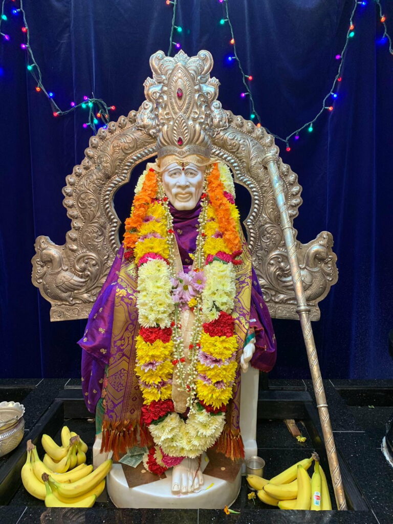 Sai Baba statue in purple-gold shawl with floral garlands on golden throne blue backdrop wallpaper