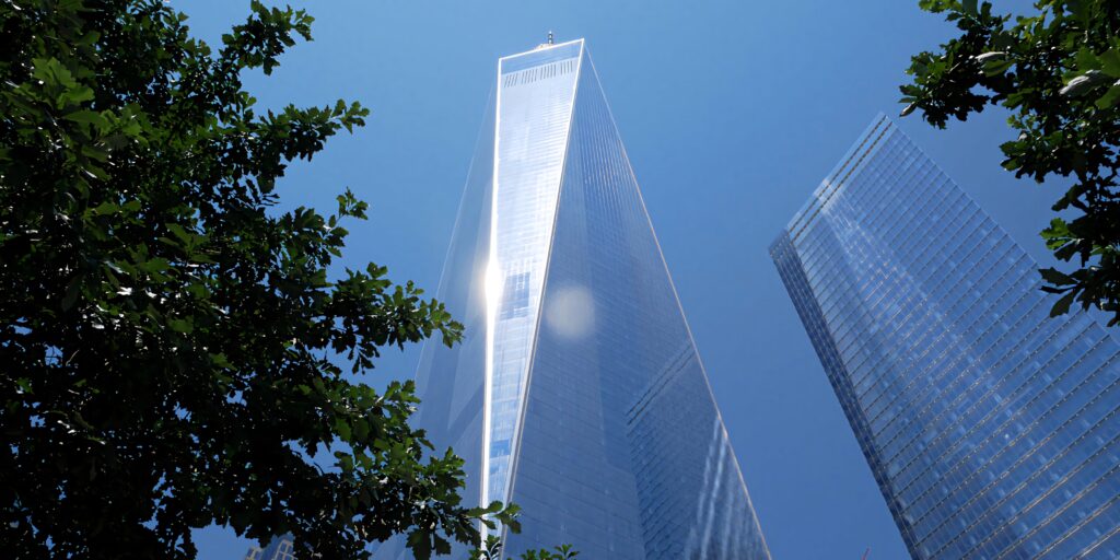 One World Trade Center towering against blue sky with green trees and modern skyscrapers background Wallpaper
