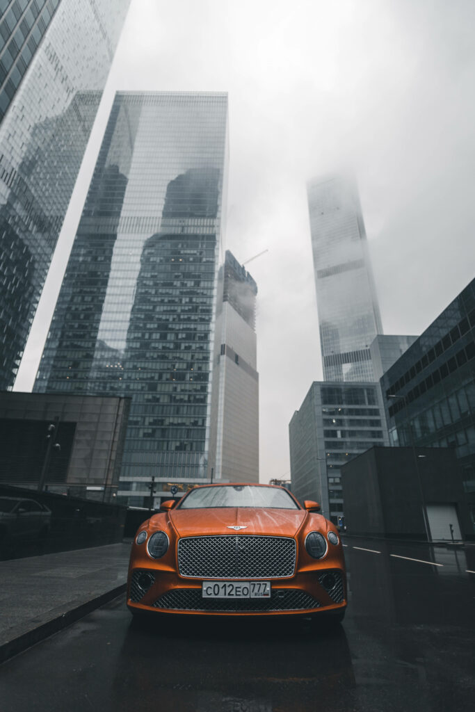 Cityscape Elegance: Orange Bentley Continental GT Graces Amidst Urban High-Rises Wallpaper