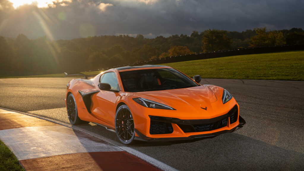 Orange Corvette sports car on racetrack under dramatic sunlight with green trees and cloudy sky Wallpaper