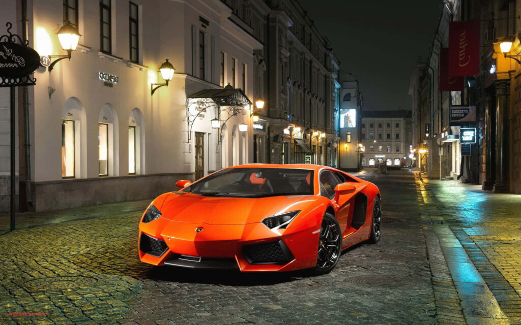 Orange Lamborghini parked on cobblestone street at night with historic buildings and soft lights Wallpaper