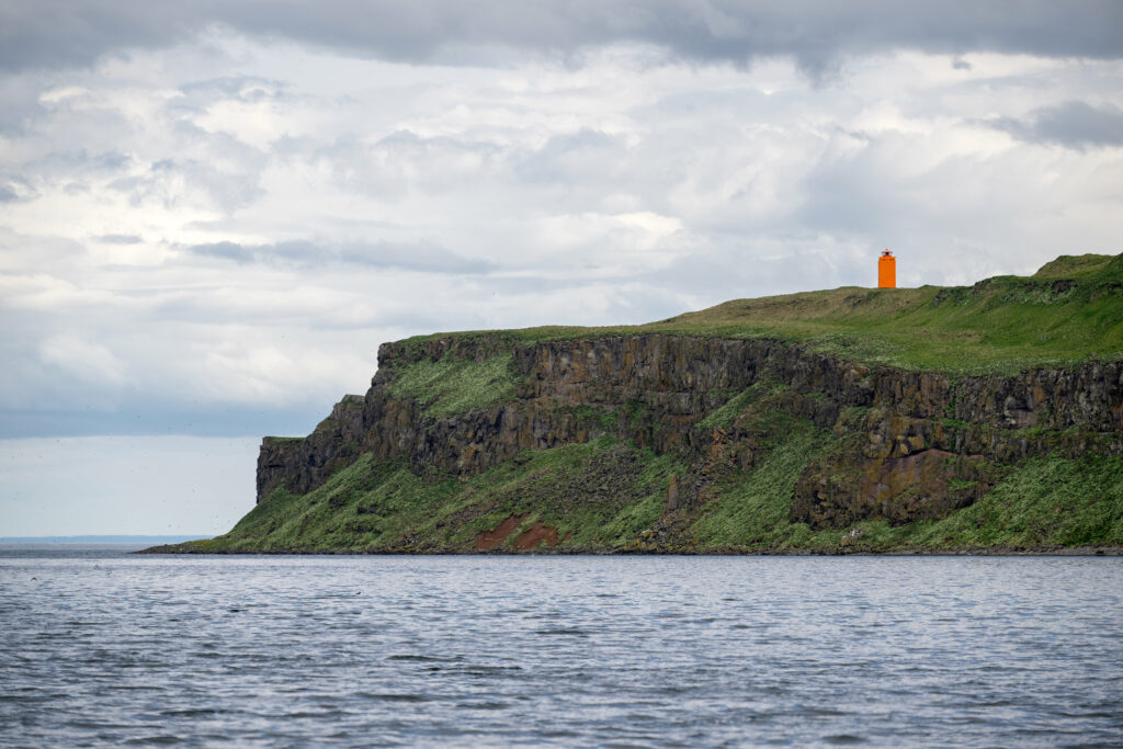Orange lighthouse on rugged grassy cliff overlooking calm blue sea with dramatic sky wallpaper