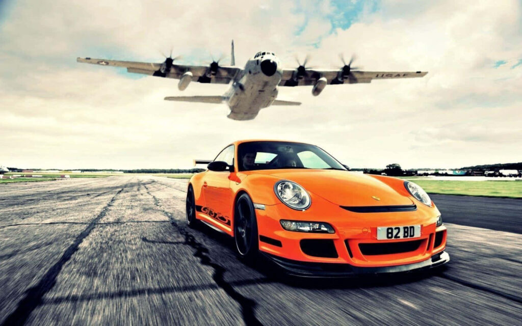 Orange Porsche 911 GT3 speeding on airstrip with low flying military transport plane wallpaper background