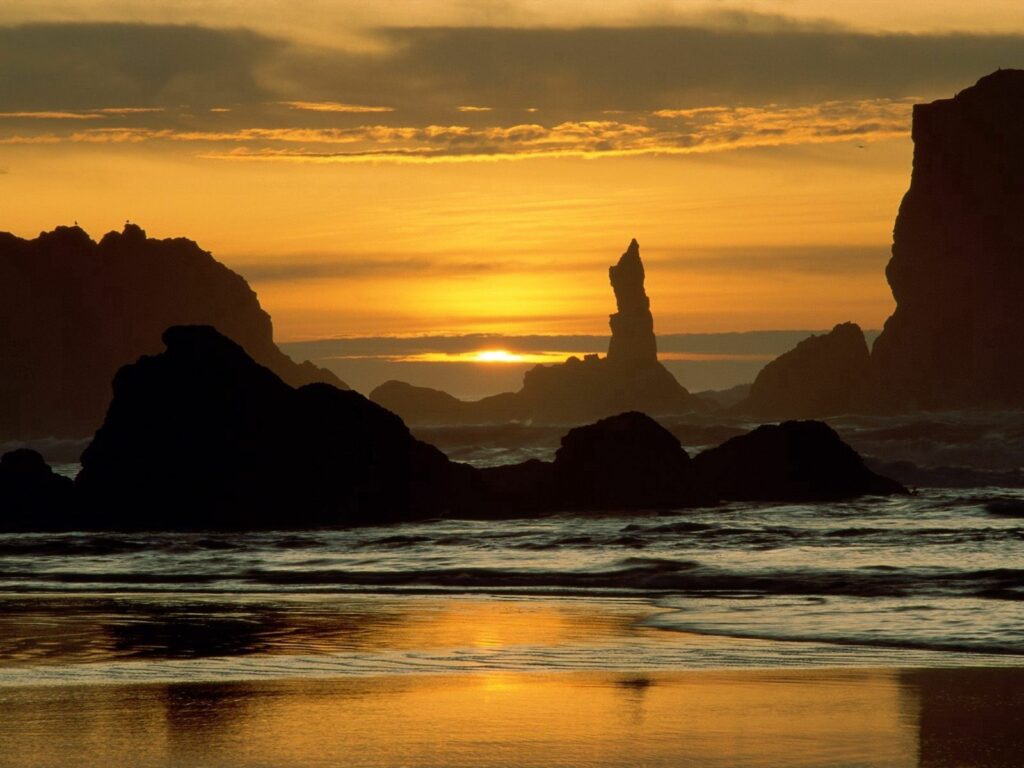 Oregon beach at sunset with towering sea rocks golden sand gentle waves and warm sky colors Wallpaper