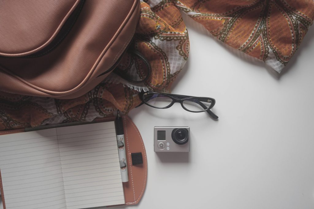 Top-down study setup with tan backpack scarf glasses notebook and silver GoPro camera wallpaper