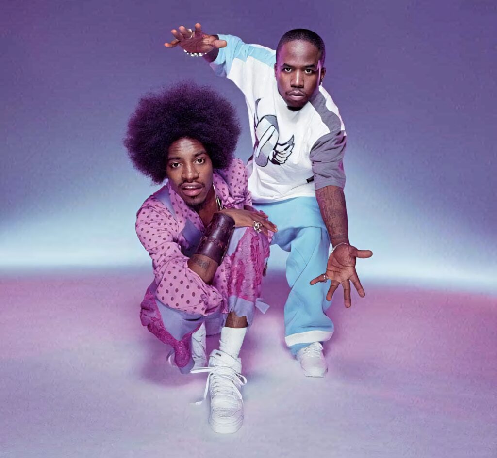 Outkast duo André 3000 and Big Boi pose dynamically with vibrant purple wallpaper background