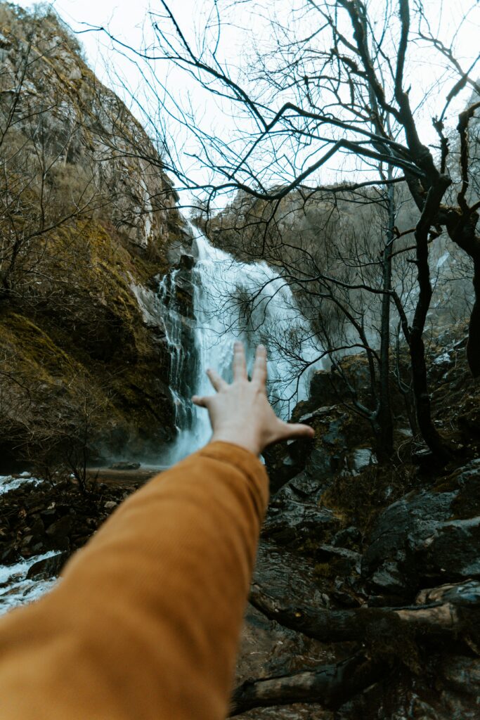 Outstretched hand reaching powerful waterfall over rugged rocks with leafless trees nature wallpaper