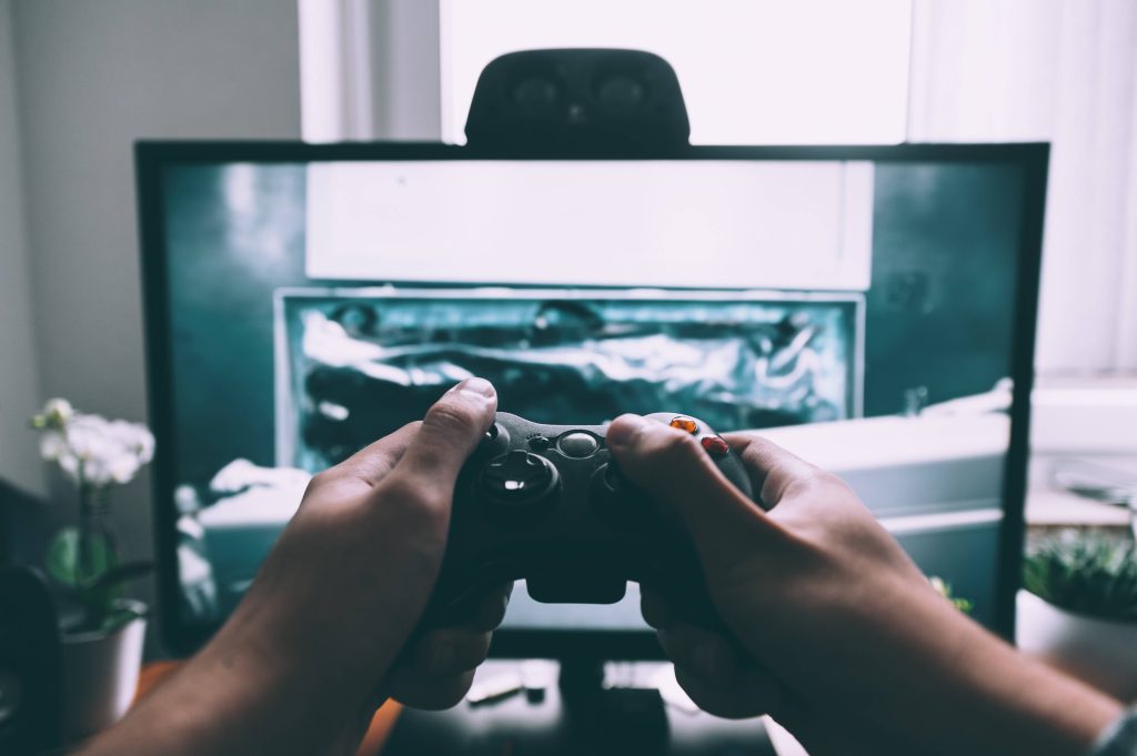 POV gaming wallpaper hands holding black controller with muted monitor and ambient desk plants
